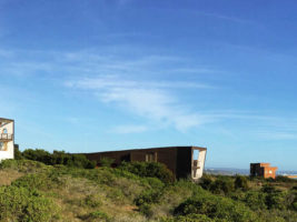 Volumen de vivienda oscuro y horizontal entre matorrales costeros, con el cielo azul de fondo